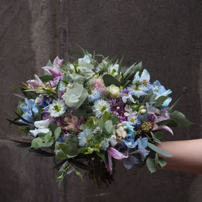 Brudebukett i pastell med blå og rosa blomster mot en steinvegg