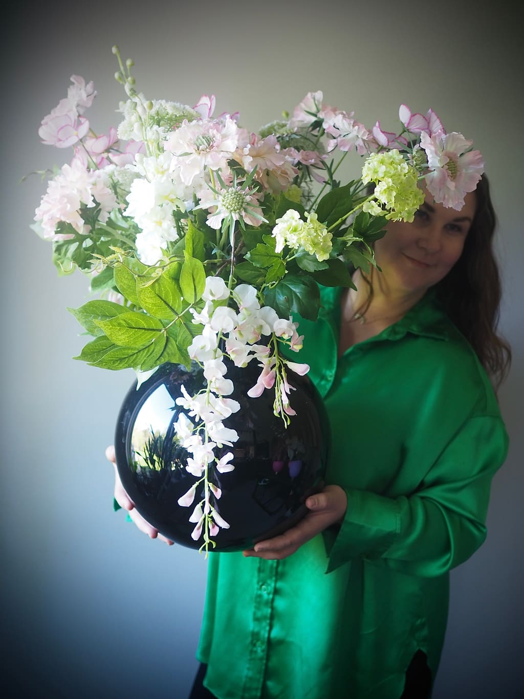 Kristin i grønn silkeskjorte holder en sort kulevase med hvite og lyserosa blomster.