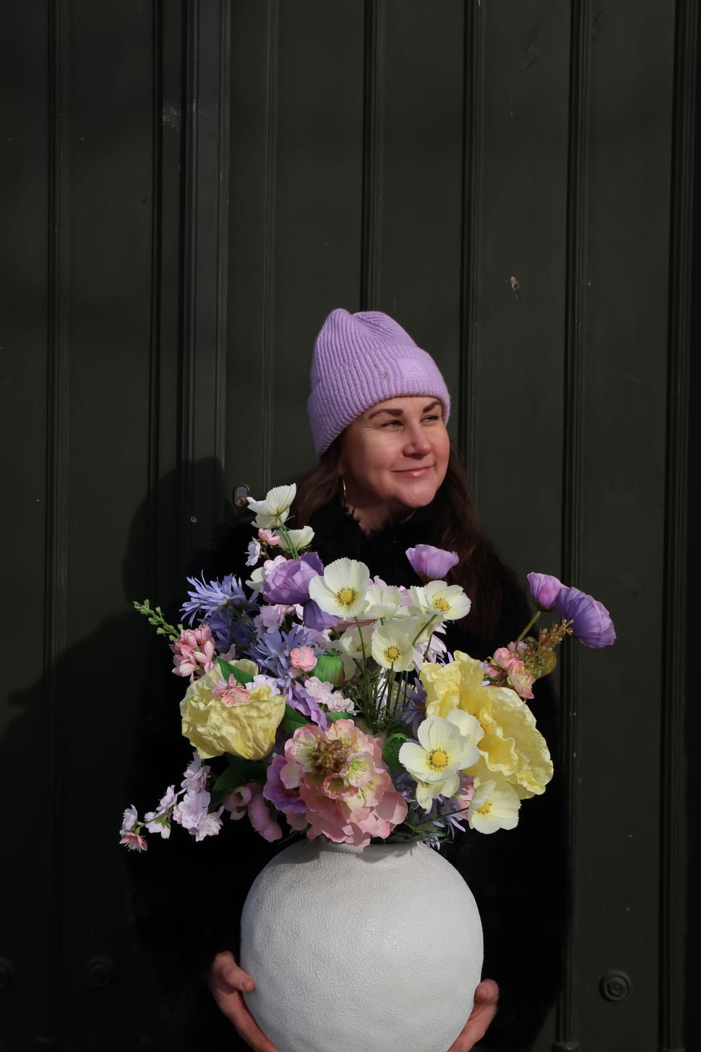 Blomsterdekoratør fra Floriss Paleet holder en vårbukett i pastellfarger foran en mørkegrønn port.